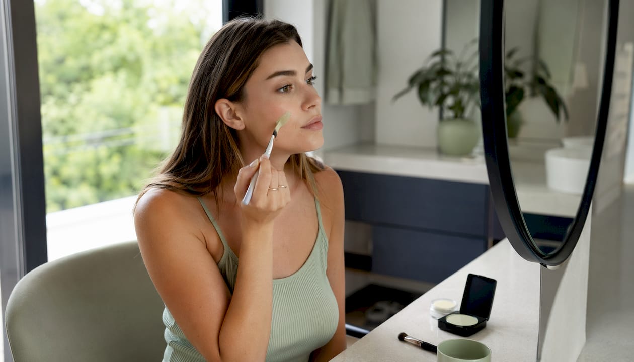 Μια γυναίκα εφαρμόζει πράσινο concealer στο πρόσωπό της για να καλύψει κοκκινίλες και ατέλειες.